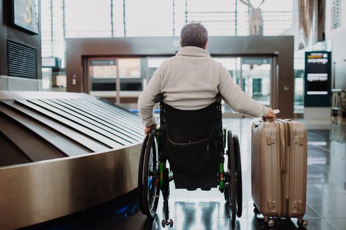 Rückenansicht eines älteren Mannes im Rollstuhl, der am Gepäckband eines Flughafens steht. Neben ihm steht ein Reisekoffer.